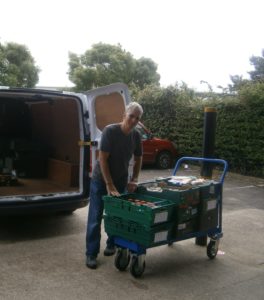 A male volunteer is standing behind a parked van which has its rear doors open. He's leaning over a trolley that has crates of donations stacked on it.