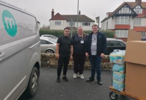 Three people stand smiling between a grey van with the Meridian Medical logo on, and a trolley piled high with cardboard boxes and packs of nappies. 