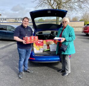 A man in foodbank uniform and a woman in a green coat standing by a car; the boot is full of donations.