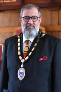 A photograph of the new Mayor of Littlehampton, Alan Butcher. Alan wears round black glasses and has grey hair and a beard. He's pictured wearing a dark suit with a red and yellow tie, and the mayoral chain of office around his neck. An oval medallion of Littlehampton's town crest hangs from the chain.